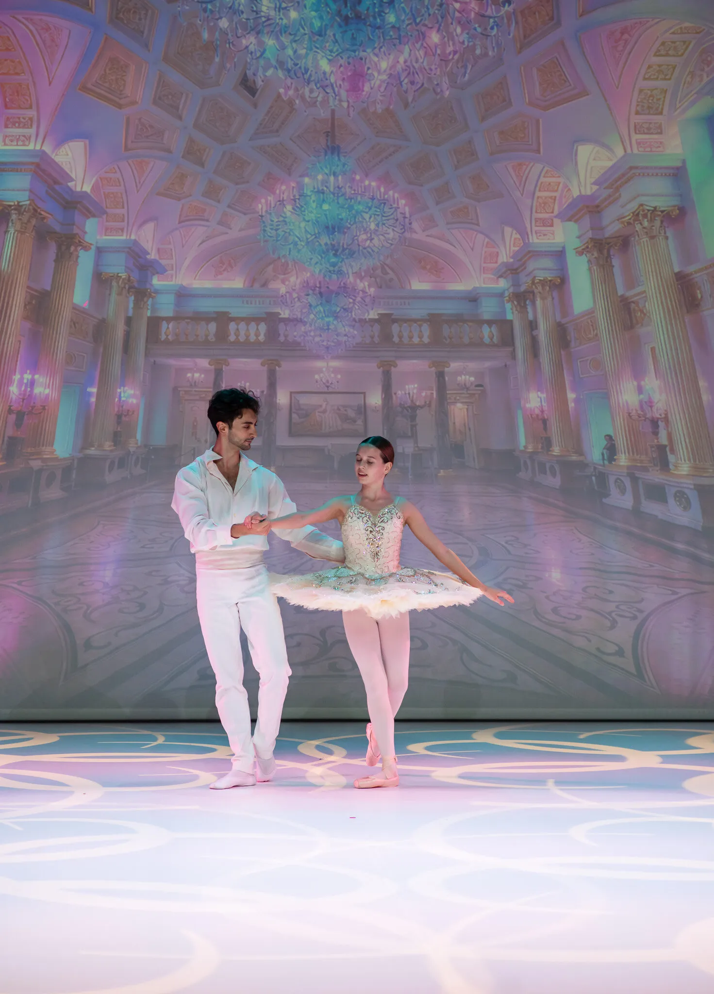 La jeune ballerine et son partenaire au centre du plateau, devant la salle de bal du palais projetée en fond.