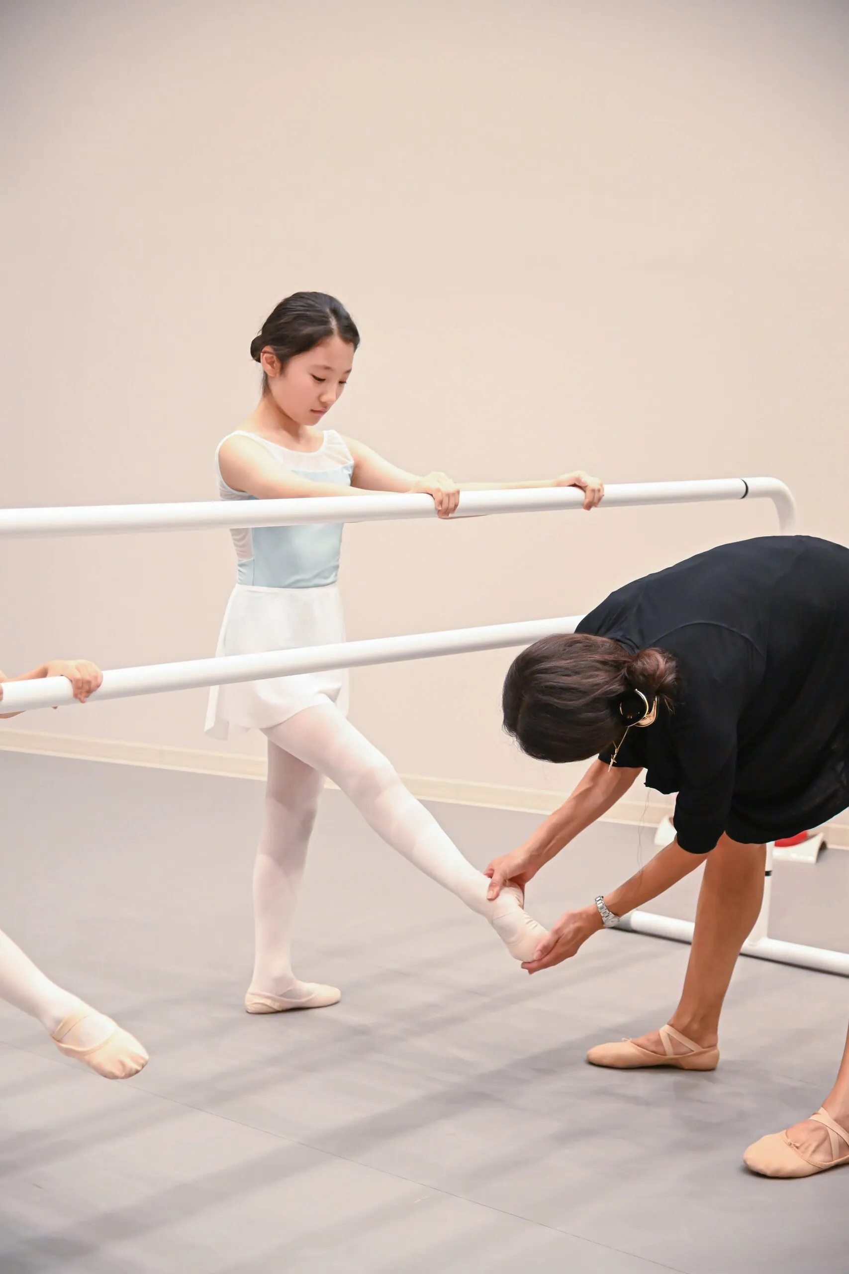 Une élève à la barre, la jambe tendue de côté, la professeure penchée qui replace son pied.