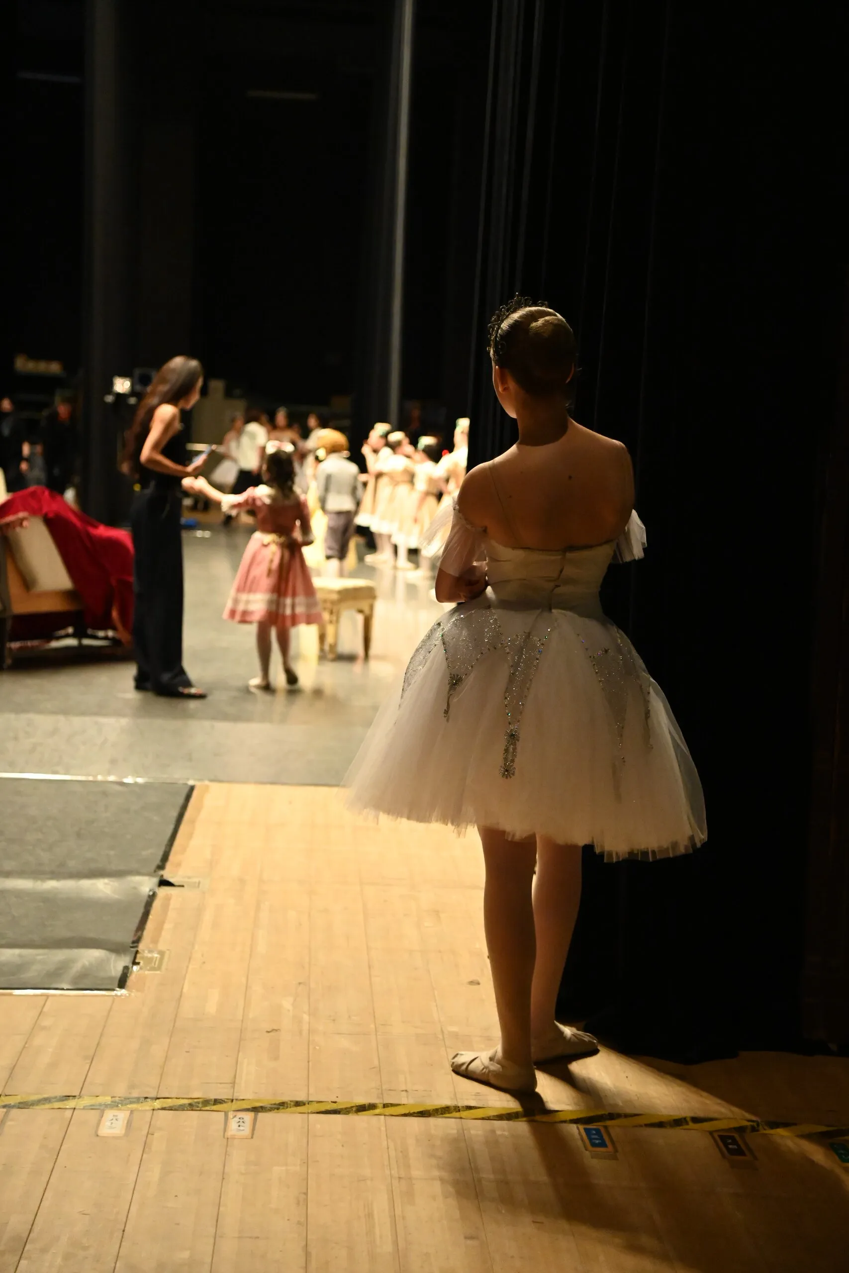 Dans les coulisses, une danseuse en tutu blanc regarde la scène.