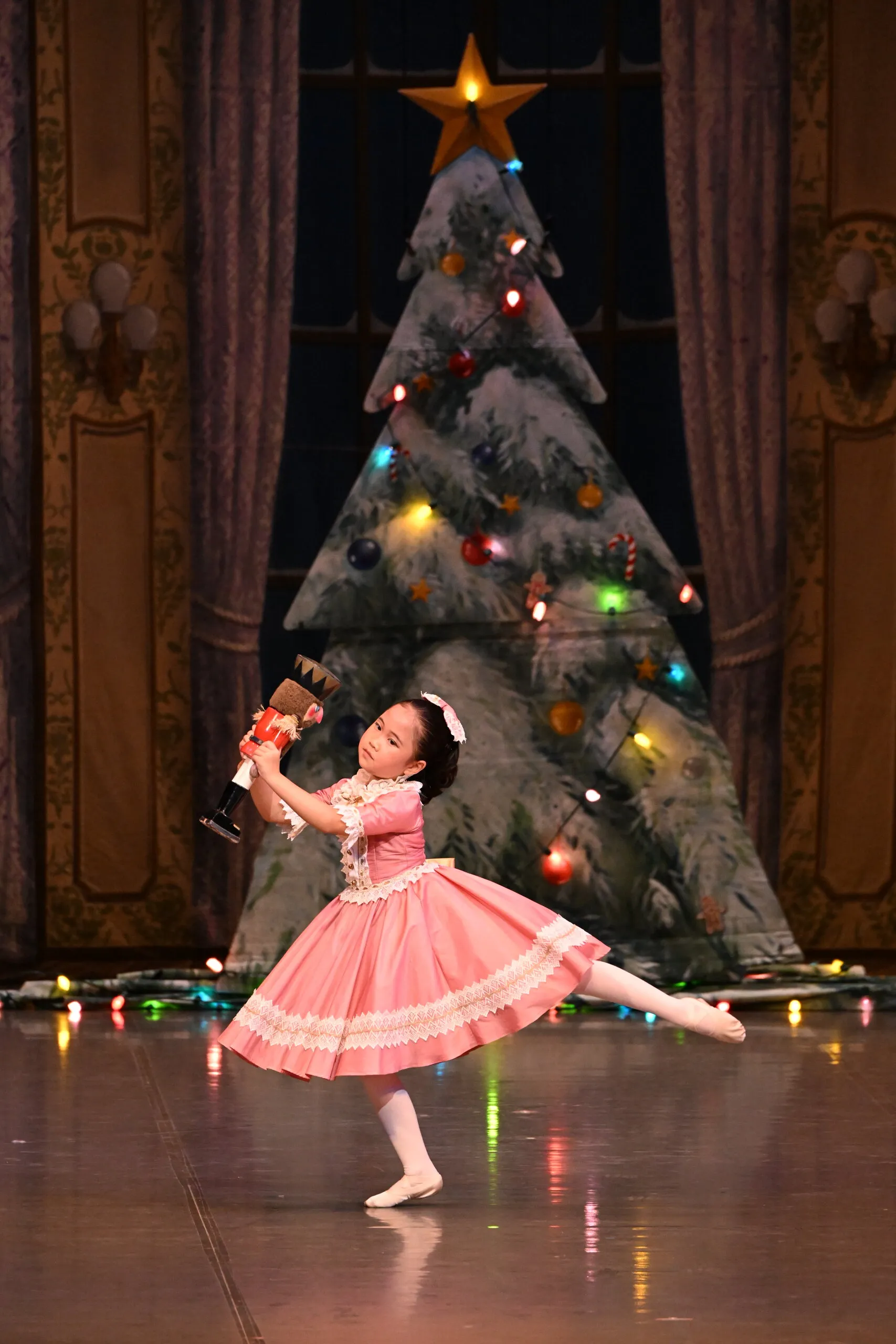 Une élève en robe rose danse avec sa poupée devant le sapin.