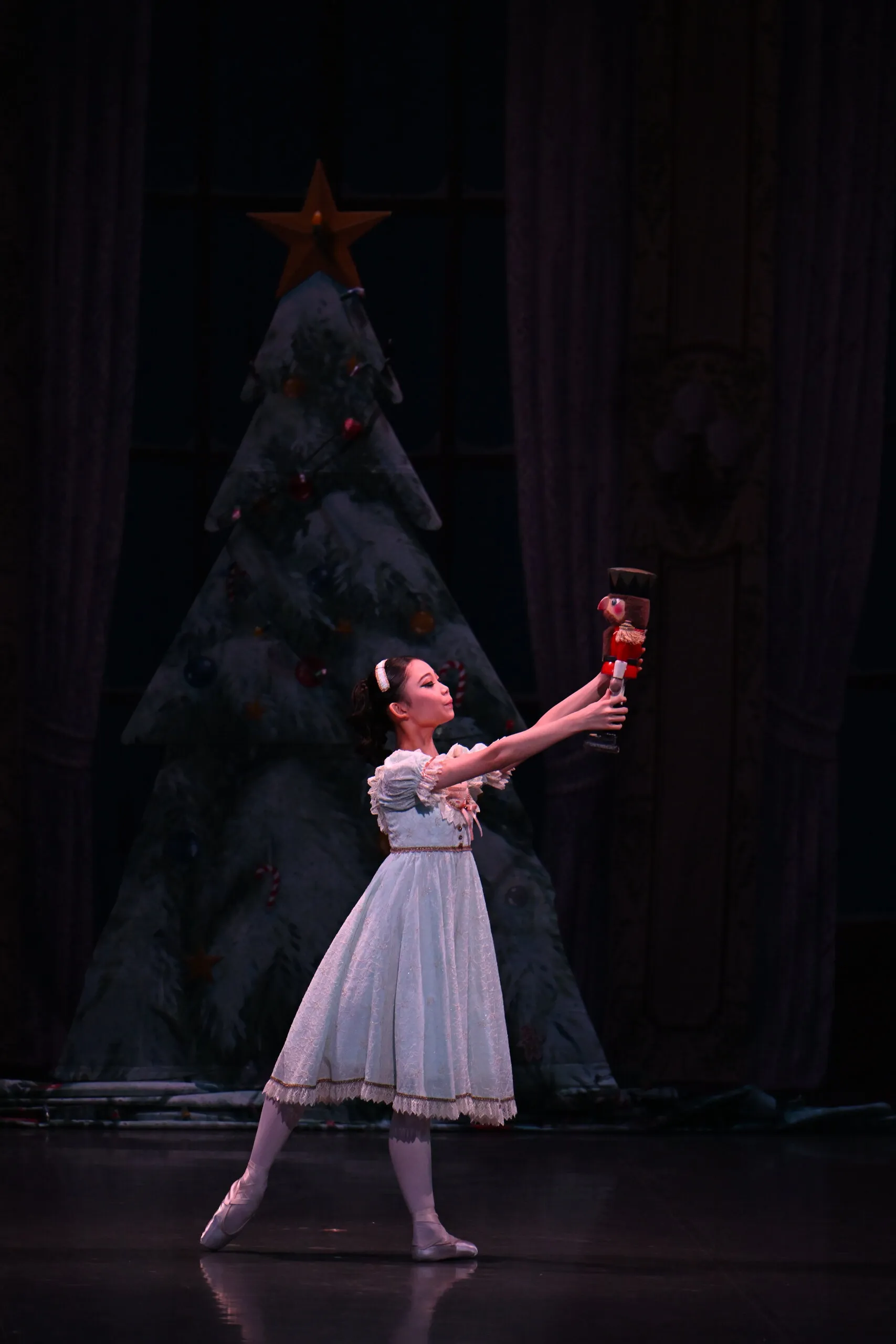 Une élève en robe claire se penche vers sa poupée, devant le sapin dans la pénombre.